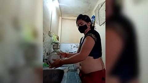 Media: Video of a woman washing dishes in a small, dimly lit kitchen with peeling walls. She wears a black mask, black top, and red pants. The image is slightly blurry.