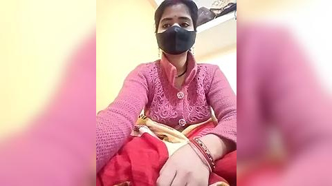 Media: Video of an Indian woman with dark skin, wearing a pink, textured cardigan, red sari, and black face mask, seated indoors, with a blurred background.