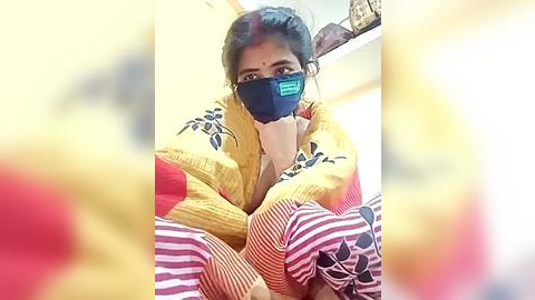 Media: Video of a South Asian woman with dark hair, wearing a yellow sweater, blue face mask, and red and white striped blanket, sitting on a bed.