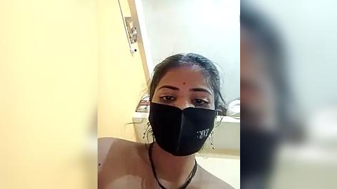 Media: Video of a young woman with medium skin tone, dark hair, and black face mask, standing in a bathroom with a yellow wall and a blurred reflection in the mirror.