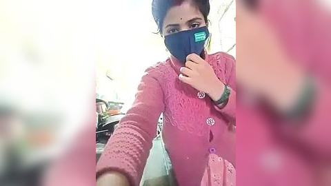 Media: A video of a young woman with medium skin tone, wearing a pink, patterned sweater and a blue face mask, indoors with a blurry background.