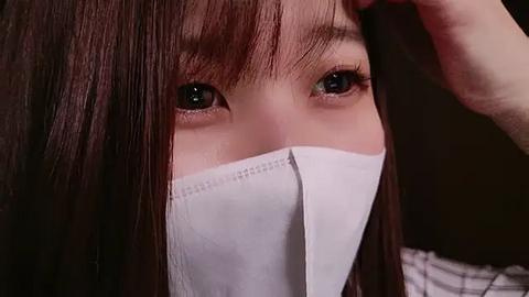 Media: A close-up video of a young Asian woman with straight, dark brown hair, wearing a white surgical mask that partially covers her face, revealing her light brown eyes and fair skin. She appears to be in a medical setting.