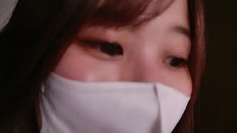 Media: A close-up video of an Asian woman with fair skin, brown hair, and expressive eyes, partially obscured by a white face mask. Her delicate features and soft skin texture are prominently visible.