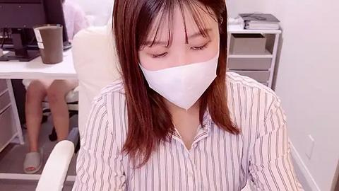 Media: Video of an Asian woman with straight, shoulder-length brown hair, wearing a white striped blouse and a white face mask, sitting at a desk in a modern office, with a computer monitor and office supplies in the background.