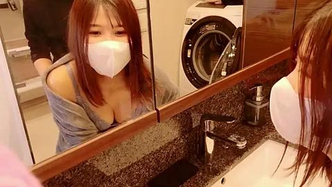 Media: Video of an Asian woman with long brown hair, wearing a white mask, leaning over a bathroom sink, looking into a mirror, revealing a cleavage in a gray top.