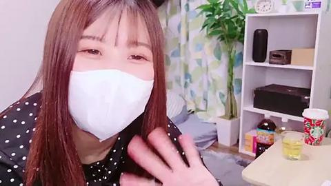 Media: Video of an Asian woman with long brown hair, wearing a white surgical mask, black dotted blouse, and looking into the camera. Background features a white shelf with plants, electronics, and a colorful mug.