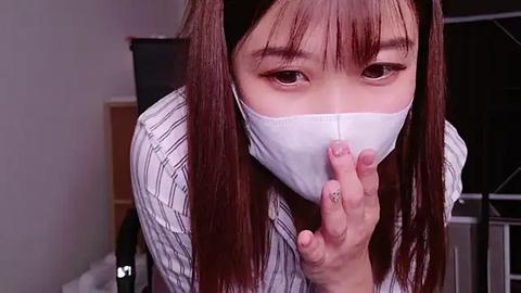 Media: Video of an Asian woman with straight, brown hair, wearing a white face mask, black-and-white striped shirt, and a gold ring on her finger, indoors with a blurred background.