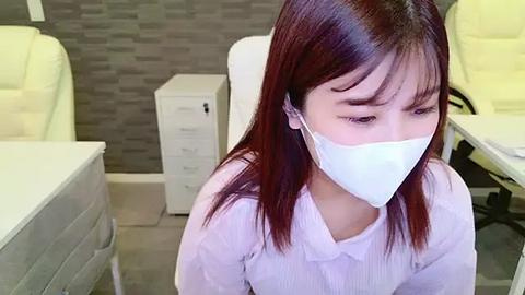 Media: Video of an Asian woman with straight, shoulder-length auburn hair, wearing a white medical mask and a white lab coat, sitting in a modern, grey-tiled medical office with white cabinets and chairs.