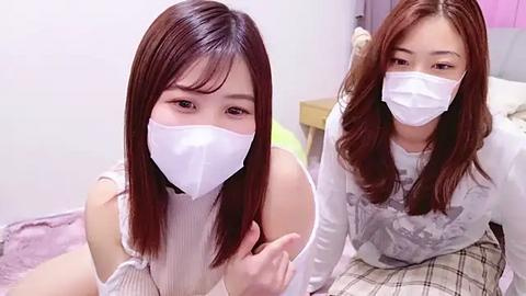 Media: Video of two Asian women with long, straight brown hair, wearing white masks, in casual outfits. One is seated on a bed, the other stands beside her. Background shows a bedroom with light-colored walls and bedding.