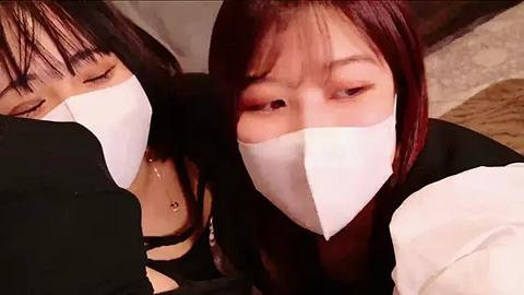 Media: Video of two Asian women with white masks, one with straight black hair, the other with straight dark brown hair, both wearing black outfits, positioned closely together, indoors with blurred background.