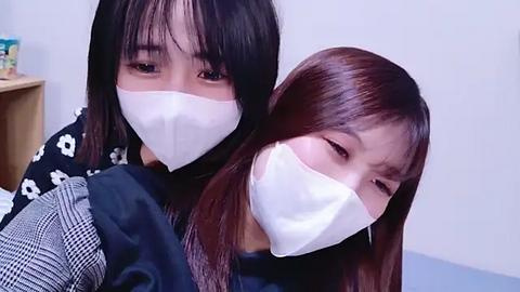 Media: Video of two Asian women with long black hair, wearing white face masks, looking down, sitting in a checkered dress and black top, against a plain white wall.