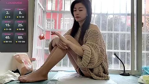 Media: Video of an Asian woman with long black hair, wearing a beige knitted sweater, sitting on a floor mat, playing a handheld game near a window with a red building in the background.