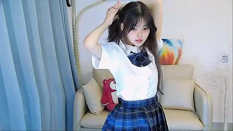 Media: Video of an Asian woman with long black hair, wearing a white blouse, plaid skirt, and red handbag, standing in a modern living room with beige armchair, colorful abstract art on the wall, and light blue curtains.