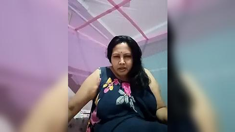 Media: Video of an overweight South Asian woman with medium skin tone, black hair, and a red bindi, wearing a sleeveless floral top, seated on a bed in a dimly lit room with pink walls and a white ceiling.