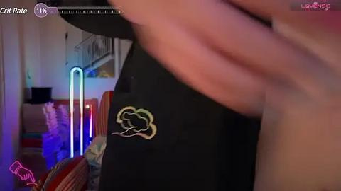 Media: Video of a close-up of a person's hand holding a black t-shirt with a yellow cloud logo, set against a dimly lit, modern room with neon lights and a bed in the background.