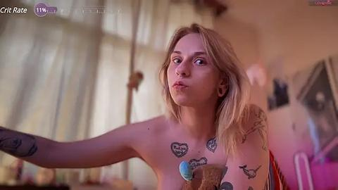 Media: A video of a topless, tattooed, fair-skinned woman with blonde hair, holding a cupcake, in a softly lit room with blurred furniture.