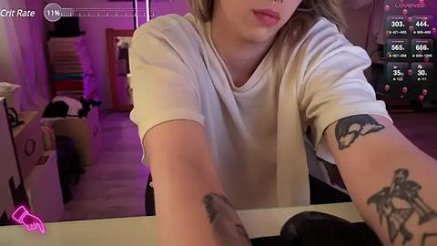 Media: A video captures a close-up of a woman with light skin, long blonde hair, and a tattoo on her forearm, seated at a desk, wearing a white t-shirt. The background shows a dimly lit room with gaming equipment and a purple light.