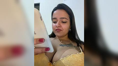 Media: A video of a Latina woman with long black hair, medium skin tone, and large breasts, wearing a yellow lace bra and a necklace, taking a selfie in a brightly lit room.