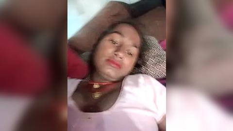 Media: A video of a woman with medium skin tone, brown hair, and a red necklace lying on a bed, looking relaxed with her arms behind her head.