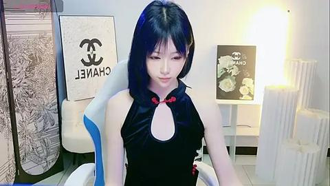 Media: Video of an East Asian woman with long black hair, wearing a black Chanel dress with a large cutout and red bow, sitting in a blue and white gaming chair, surrounded by minimalist decor.