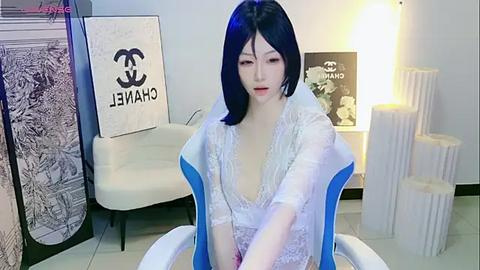 Media: Video of an Asian woman with pale skin and shoulder-length black hair, wearing a white lace dress, sitting in a modern room with a white chair, Chanel art, and tall white candles.