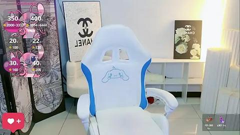 Media: Video of a modern white gaming chair with blue accents, placed in a minimalist room with white walls, a white desk, and a large floral poster. The room has a clean, sleek design.