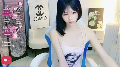 Media: Video of an East Asian woman with fair skin and straight black hair, wearing a white tank top with a graphic, sitting in a modern living room with a CHANEL sign and a pink My Little Pony plush toy.