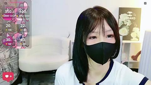 Media: Video of an East Asian woman with straight black hair, wearing a black face mask, white T-shirt, and sitting indoors. The background includes a white couch and a bookshelf with white flowers and a \"CANADA\" sign.