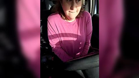 Media: Video of a woman in a car, wearing a pink long-sleeve shirt, smiling, with sunlit window reflections creating a surreal, almost dreamlike atmosphere.