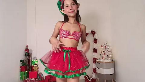 Media: Video of a young Asian woman in a festive red and green elf costume, posing with a playful expression. Background features Christmas decorations, including wrapped gifts and a decorated tree.