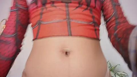 Media: Video of a person wearing a red and black Spider-Man costume with long sleeves, showing their midriff and belly button. Background is a plain white wall with blurred objects.