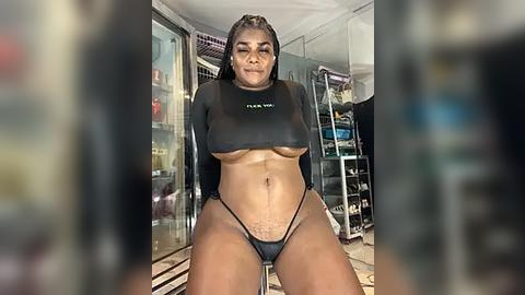 Media: Video of a young Black woman with braids, wearing a black crop top and thong, posing in a modern, brightly lit room with glass walls, shelves, and a staircase.