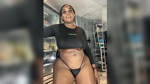 Media: Video of a confident, dark-skinned woman with a fit physique wearing a black crop top and thong, standing in a modern kitchen with stainless steel appliances and glass cabinets.