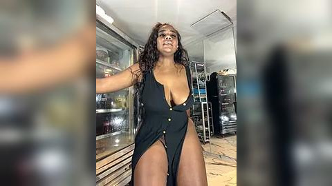 Media: Video of a confident, dark-skinned woman with long, wavy hair, wearing a black, plunging one-piece swimsuit, posing in a modern, glass-walled room with blurred background.