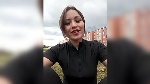Media: Video of a smiling woman with dark hair, wearing a black shirt, sitting outdoors on grass with a red-brick building in the background.