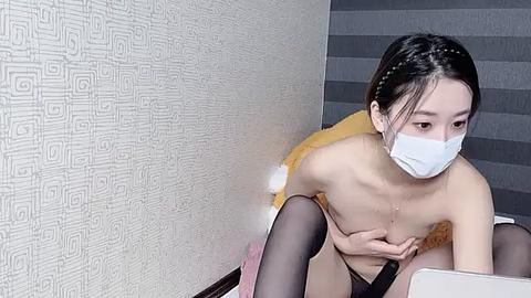 Media: Video of an East Asian woman with short black hair, wearing a white face mask and sheer black thigh-high stockings, crouching topless, revealing small breasts. Background features textured wallpaper and a striped cushion.