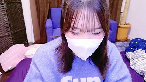 Media: A video of an Asian woman with long brown hair and white face mask, wearing a blue sweatshirt, sitting on a purple bed, surrounded by clothes, in a cluttered bedroom.