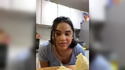 Media: Video of a woman with dark hair, wearing a gray t-shirt, seated at a kitchen table, smiling, with blurred background featuring white cabinets, a refrigerator, and kitchen utensils.