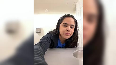 Media: Video of a young woman with long dark hair in a blue top, leaning over a sink in a white-tiled bathroom. The image is slightly blurred.