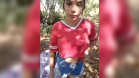 Media: Video of a young woman with light skin and brown hair, wearing a red crop top and denim shorts, standing in a wooded area with sunlit leaves.