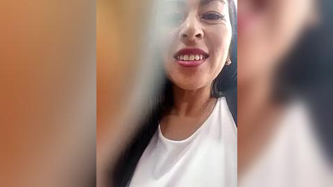 Media: Video of a smiling woman with medium brown skin, wearing a white top, taken through a blurry, multi-colored window.