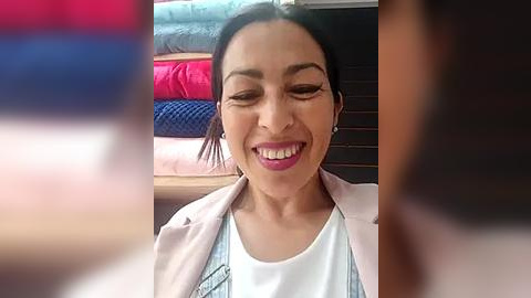 Media: Video of a joyful woman with medium skin tone, dark hair, and pink lipstick, wearing a light pink jacket over a white shirt, sitting in a room with folded clothes in the background.