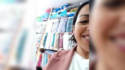 Media: Video of a smiling Asian woman with medium skin tone and dark hair, wearing a pink blouse, standing in front of a colorful, neatly organized rack of fabric rolls in a sewing store.