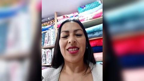 Media: Video of a smiling woman with long black hair, wearing a light-colored blazer, in a vibrant fabric store with colorful, neatly stacked rolls of fabric in the background.