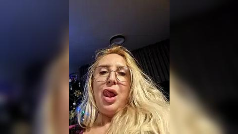 Media: Video of a young woman with long blonde hair, wearing glasses, sticking out her tongue. Background features a dimly lit room with a ceiling light and a blurred, colorful object in the distance.