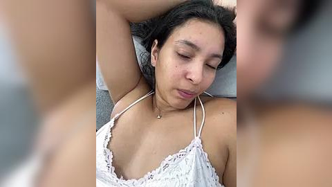 Media: Video of a young woman with medium skin tone, long dark hair, lying on her back, wearing a white lace camisole, eyes closed, arms raised, relaxed expression.