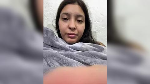 Media: Video of a young woman with long black hair, medium skin tone, and light makeup, wrapped in a gray blanket, indoors against a white textured wall.