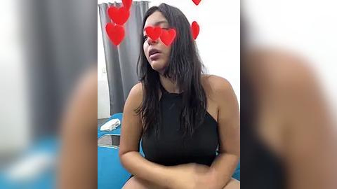 Media: Video of a woman with long black hair, wearing a black tank top, heart emojis over her eyes, sitting on a blue couch in a room with gray curtains and a white wall.