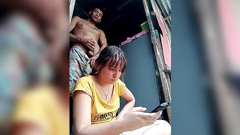 Media: A video showing a shirtless, muscular man with a tattoo on his chest, wearing plaid shorts, standing behind a young woman with shoulder-length brown hair in a yellow shirt, who's focused on her smartphone.