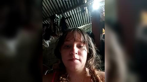 Media: Video of a young, fair-skinned woman with shoulder-length, wet, brown hair, looking directly at the camera. She is inside a dimly lit, rustic room with a corrugated metal roof, wooden walls, and a few hanging objects.